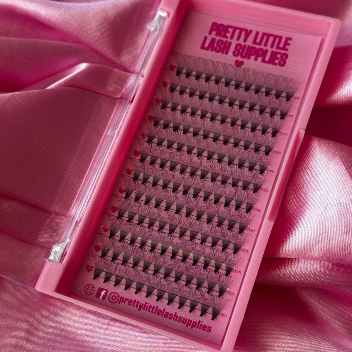 Pink lash extension tray containing black individual lashes in varying lengths on a satin backdrop.