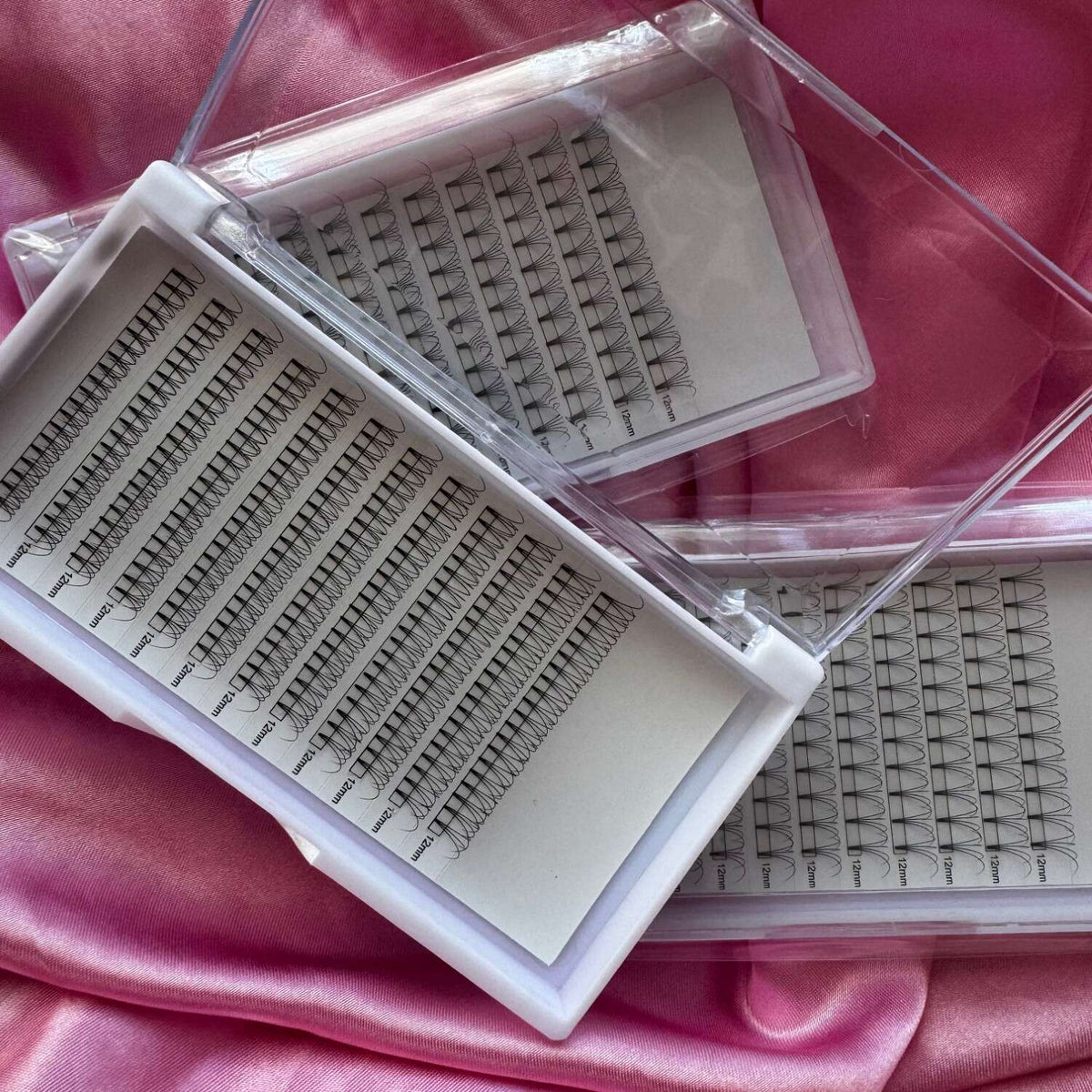 Clear storage cases with black volume eyelash extensions, featuring varied lengths, on satin pink fabric background.