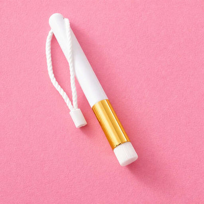 White makeup brush with gold accents and a braided rope on a pink background. Ideal for precision application.