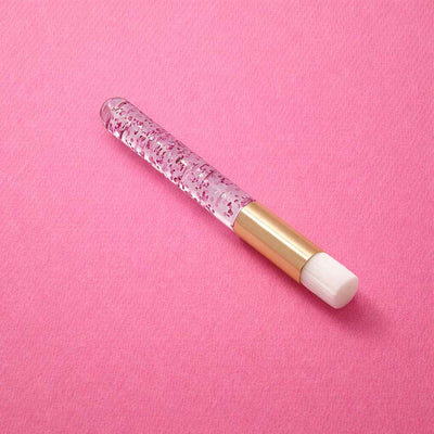 Clear acrylic makeup brush cleaner with gold accents, featuring pink glitter and a white applicator tip on a pink background.
