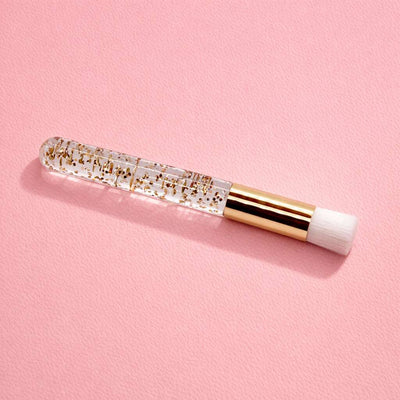Clear cosmetic brush with gold glitter, white bristles, and a chic gold handle over a soft pink background.