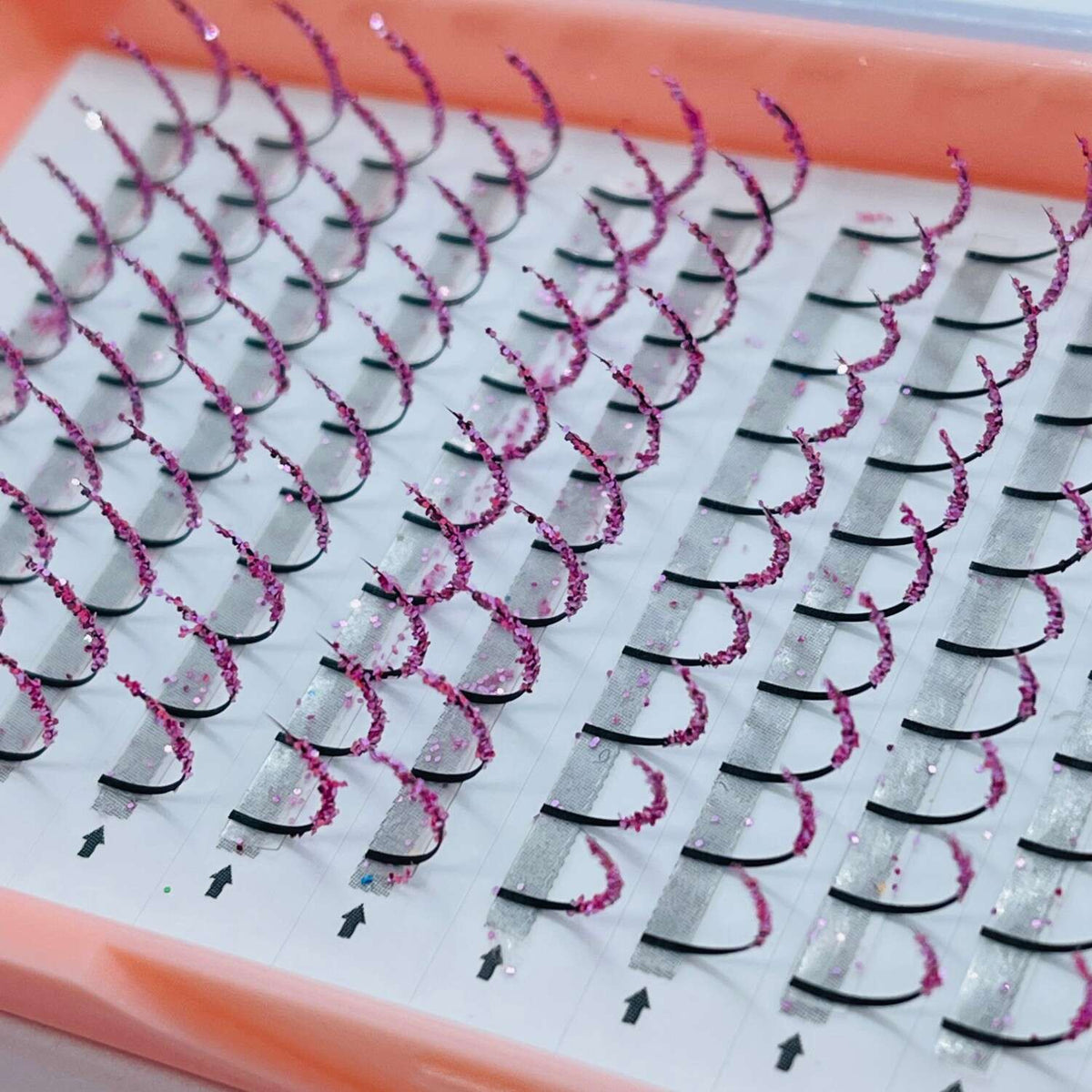 Pink glitter eyelash extensions with black bases in a clear tray, perfect for glam makeup looks.