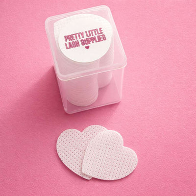 White lash supply pads in heart shapes, packaged in a clear container with a colorful label, set against a pink background.