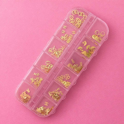 Gold metal charms in a clear organizer, featuring various shapes like flowers and logos on a pink background.