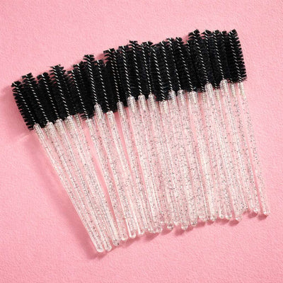 Clear glitter eyelash and brow wands with black bristles on a pink background, perfect for makeup application.