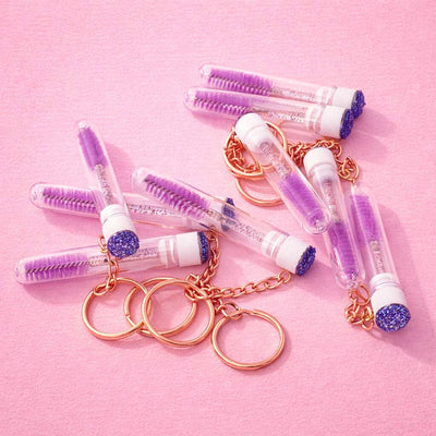 Purple mascara wands in clear tubes with glittery caps, on rose gold keychains, perfect for stylish makeup enthusiasts.