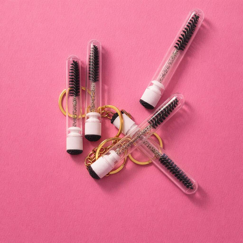 Set of clear plastic mascara wands with black bristles, featuring gold keyring attachments, on a pink background.