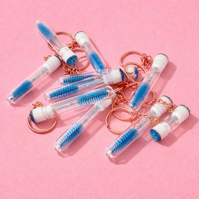 Blue glitter keychain brushes with transparent tubes and rose gold accents on a pink background.