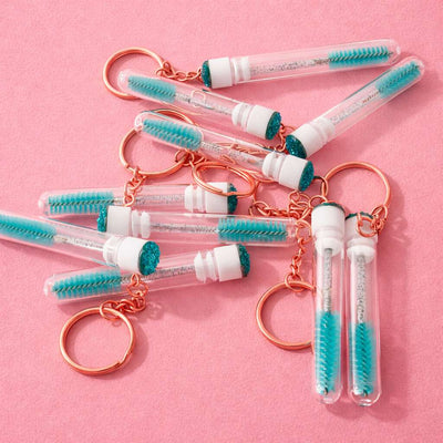 Clear acrylic keychain mini brushes with teal bristles and rose gold accents on a pink background.