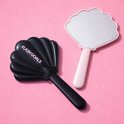 Black shell-shaped mirror with "#LASHGOALS" text and a pink hand-held mirror on a pink background.