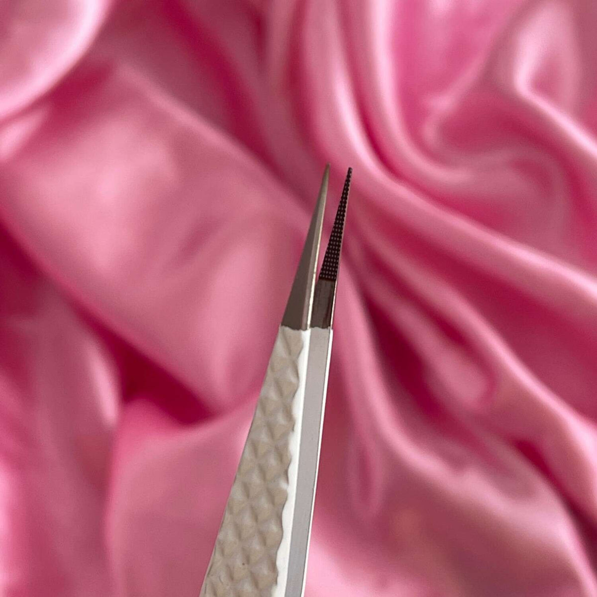 Silver stainless steel tweezers with textured white handle, set against a soft pink satin background.